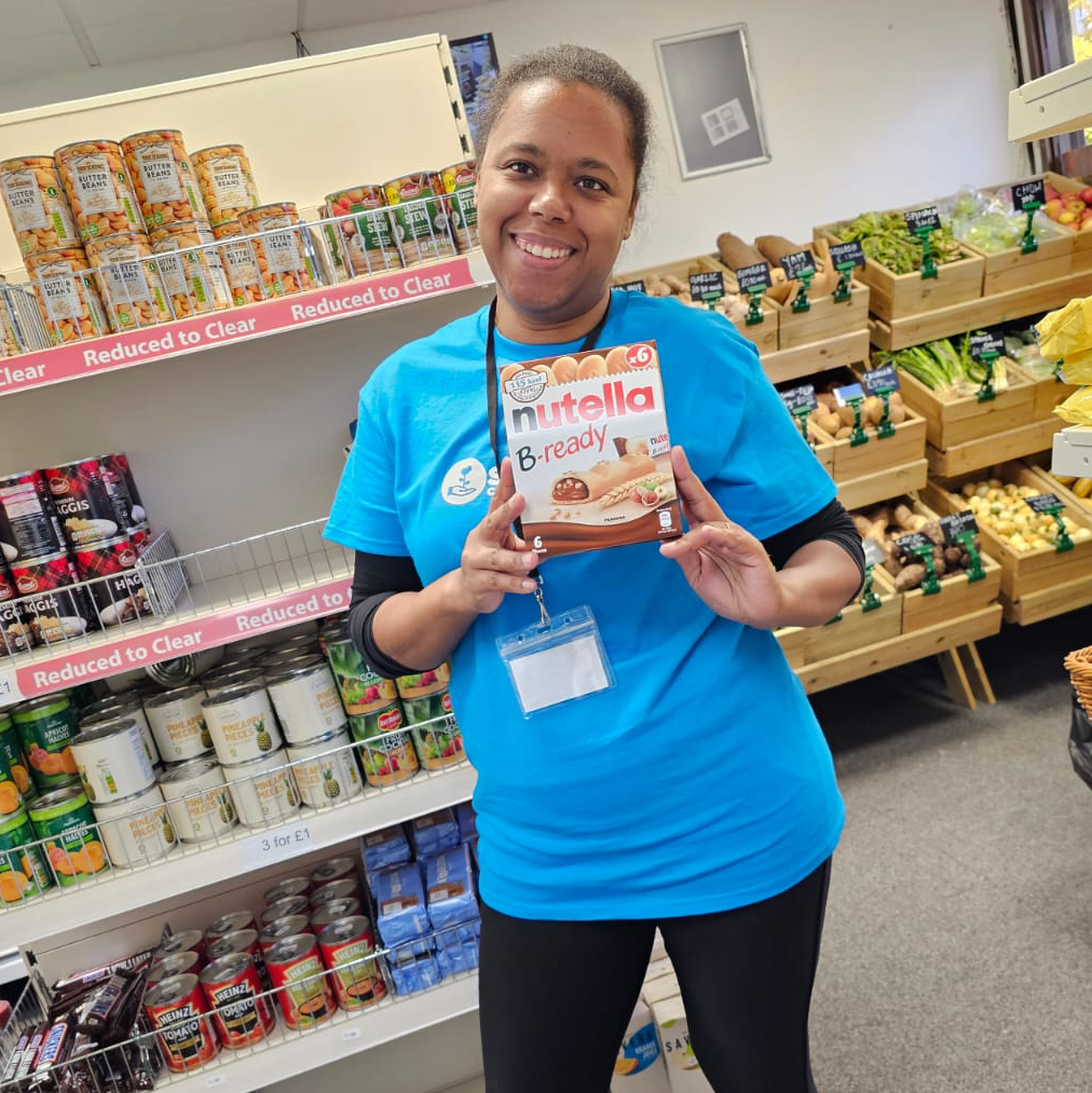 A woman in a blue SCH Tshirt stands smiling in front of supermarket shelves, holding a box of Nutella BeReady bars