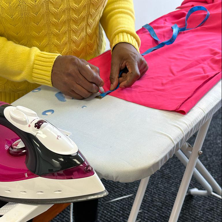 woman in a yellow jumper measuring pink material on an ironing board