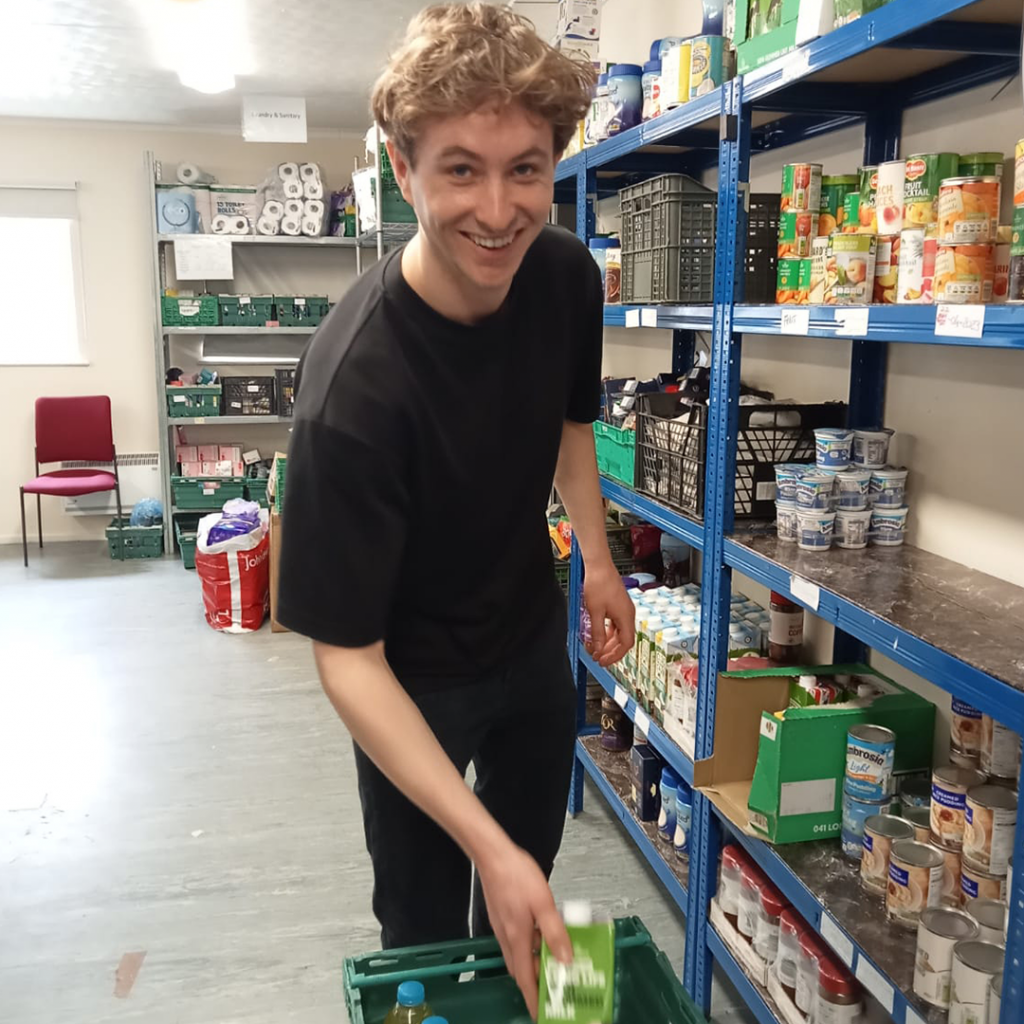 A young man volunteering in food bank smiles at the camera while putting a carton of juice into a basket