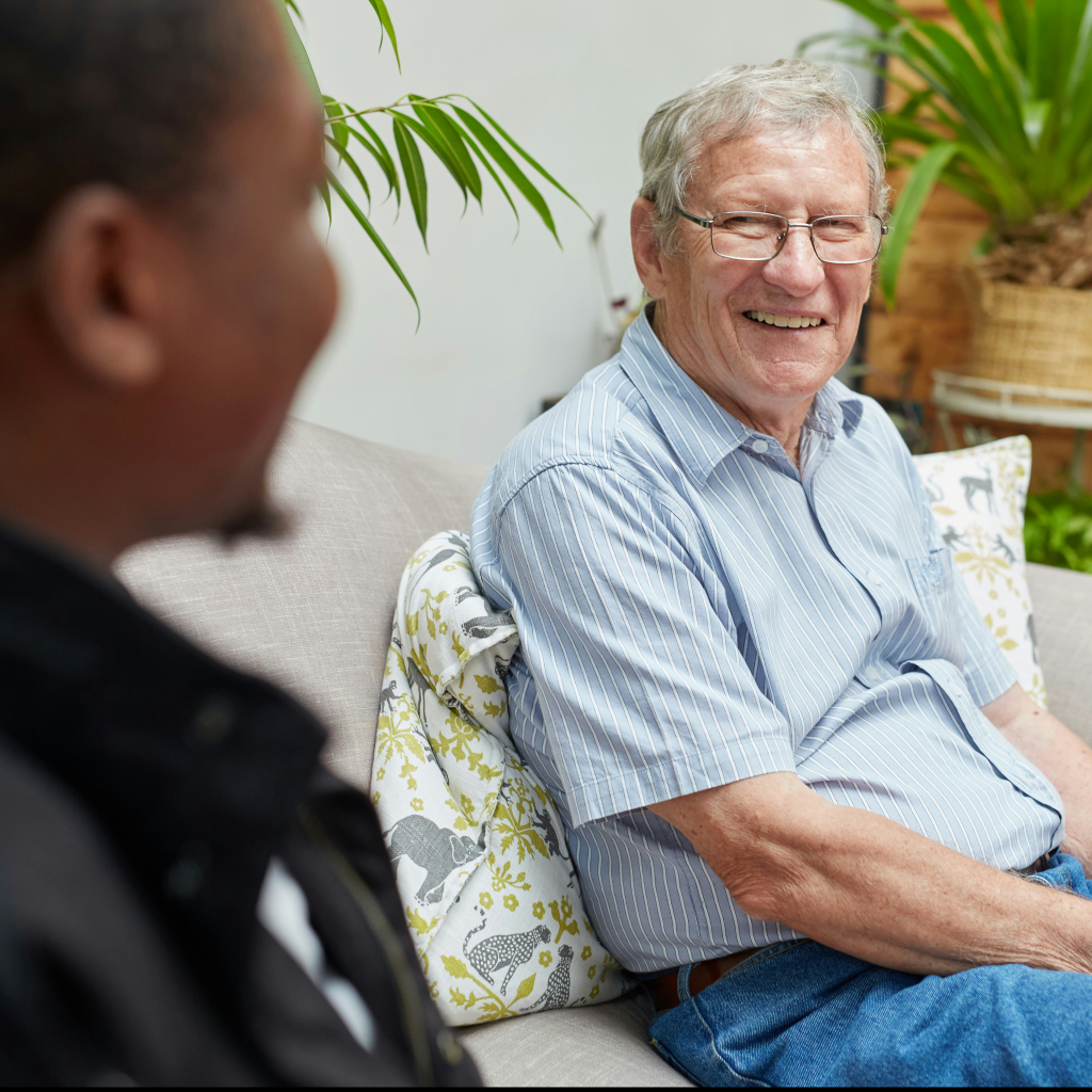 An elderly man with gray hair and glasses smiles while sitting on a couch, engaged in conversation with another person in a cozy living room with plants.