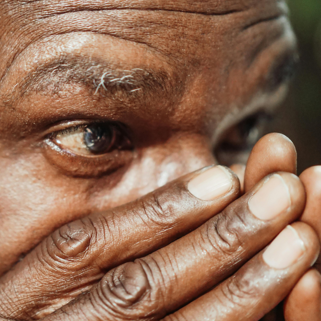 Close-up of an older man’s eye and face, with his hand partially covering his mouth, showing detailed skin texture and a thoughtful expression.