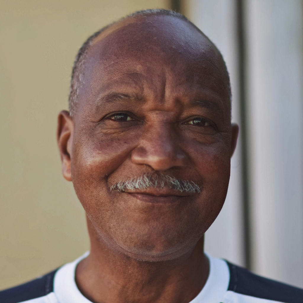 Portrait of an older man with a mustache smiling gently at the camera against a neutral background.