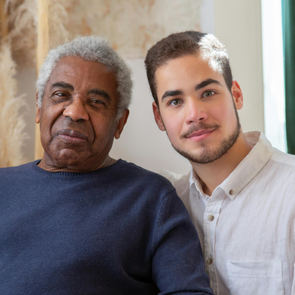 An older man and a younger man sit closely together, both looking at the camera with calm expressions in a warmly lit indoor setting.
