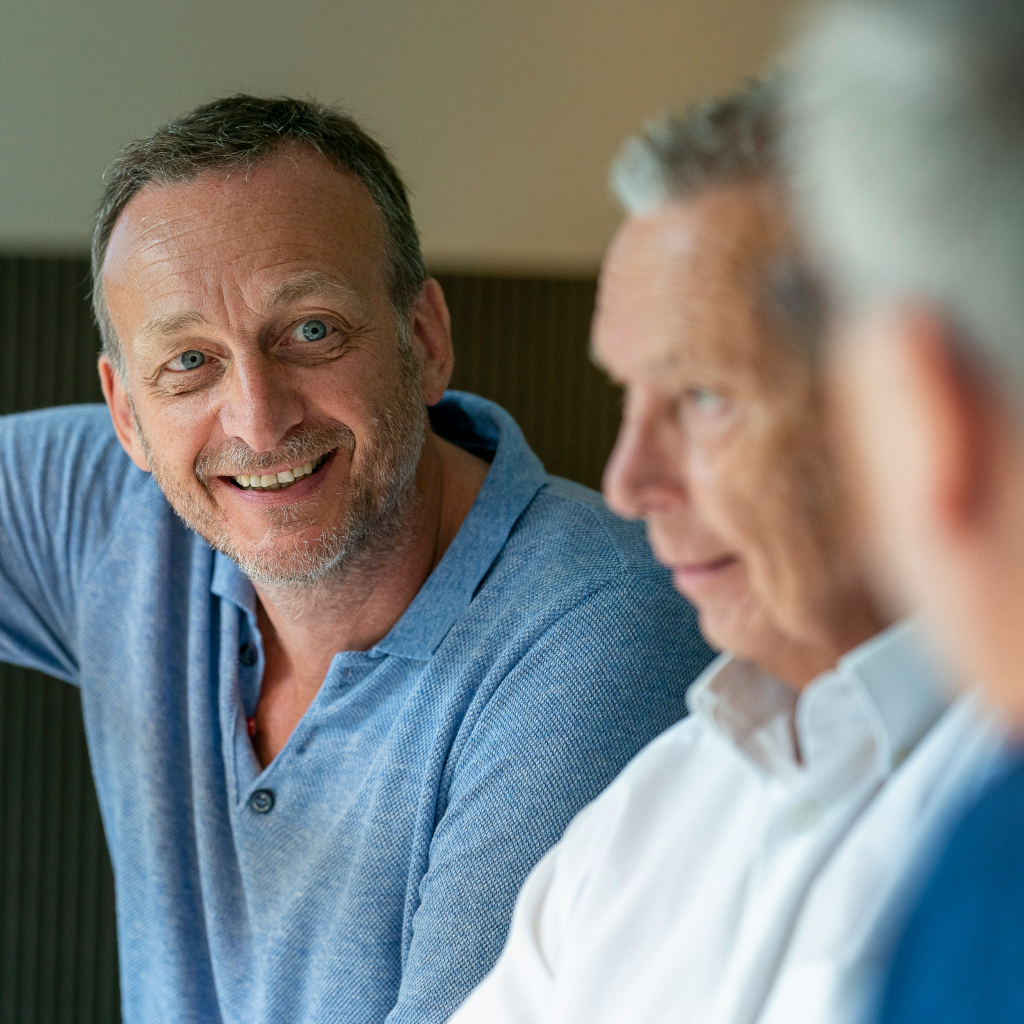 A middle-aged man smiles warmly while talking to two other people, slightly out of focus, in a casual indoor environment.